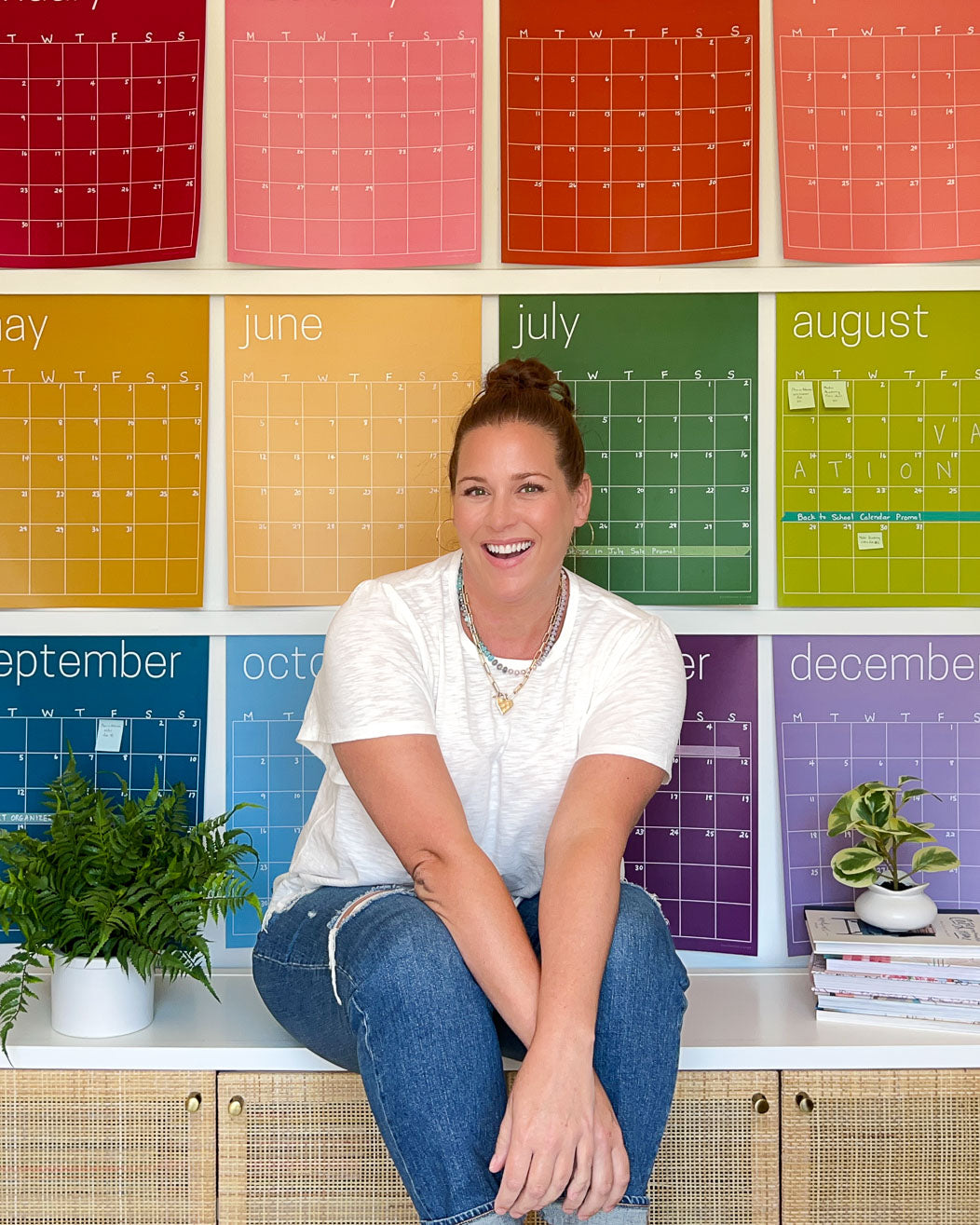 photo of Tasha Agruso sitting on console table in front of large rainbow wall calendar from Kaleidoscope Living