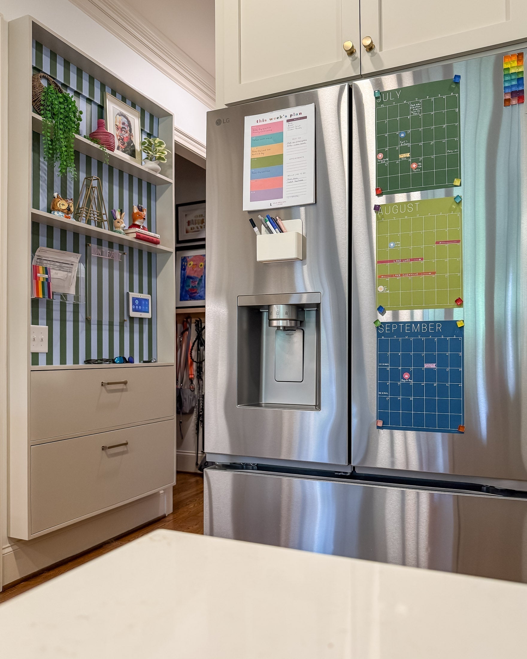 A stainless steel French-door refrigerator in a kitchen setting, with colorful monthly calendar hanging on it. To the left is a hanging shelving unit backed with striped blue-and-green wallpaper and featuring a family command and decorative items.