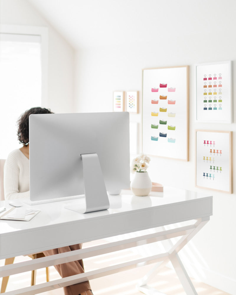 A white office with a women sitting at a desk playing several framed office themed art prints with ombre rainbow hues and a white background.