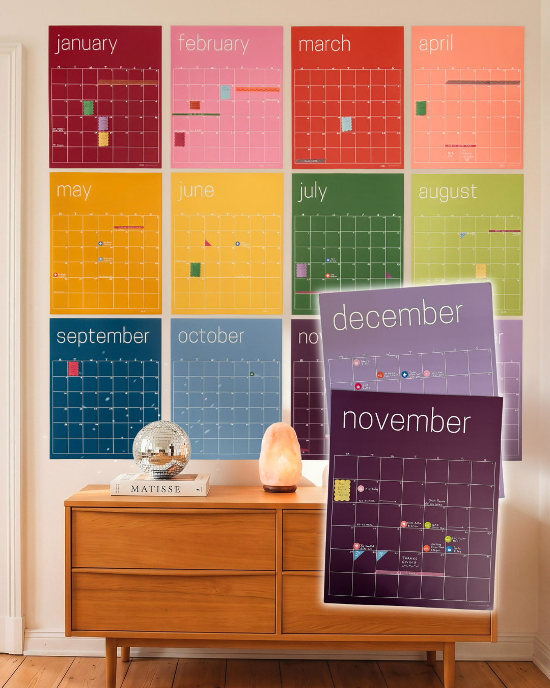 Twelve large, brightly colored reusable monthly wall calendars displayed in a rainbow arrangement above a wooden dresser with decorative accessories with November and December highlighted in the foreground.