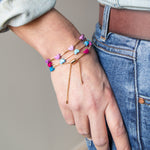 Hand wearing three colorful bracelets with pink, blue and purple hearts and a gold adjustable chain.