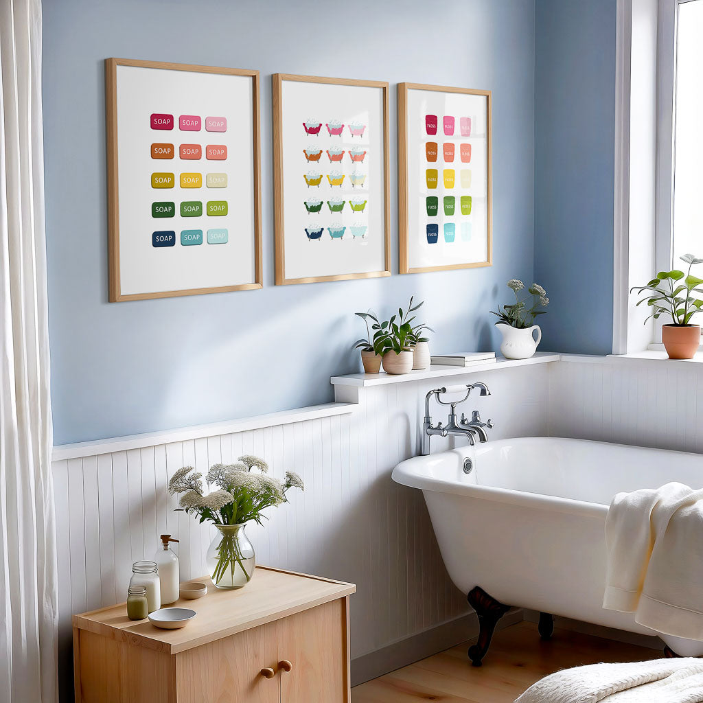 A set of three colorful art prints with white backgrounds, displayed above a bathtub and a wooden cabinet in a bathroom, displaying picture of soap, bathtubs and floss in ombre rainbow hues.