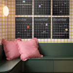 Six black monthly calendar posters from July to December hanging on a wall with pink, green and white checkered wallpaper above a dark green built-in bench with pink and red striped pillows.