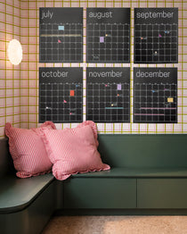 Six black monthly calendar posters from July to December hanging on a wall with pink, green and white checkered wallpaper above a dark green built-in bench with pink and red striped pillows.