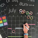 Erasable black calendar with colorful washi tape and sticky notes with woman's hand writing on it in white chalk marker