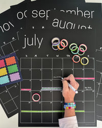Erasable black wall calendar with colorful washi tape and sticky notes with a woman's hand pictured writing on it with white chalk marker