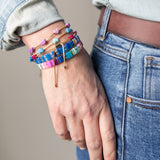 Hand wearing a stack of colorful bracelets including ones with enamel hearts and other with resin tiles.