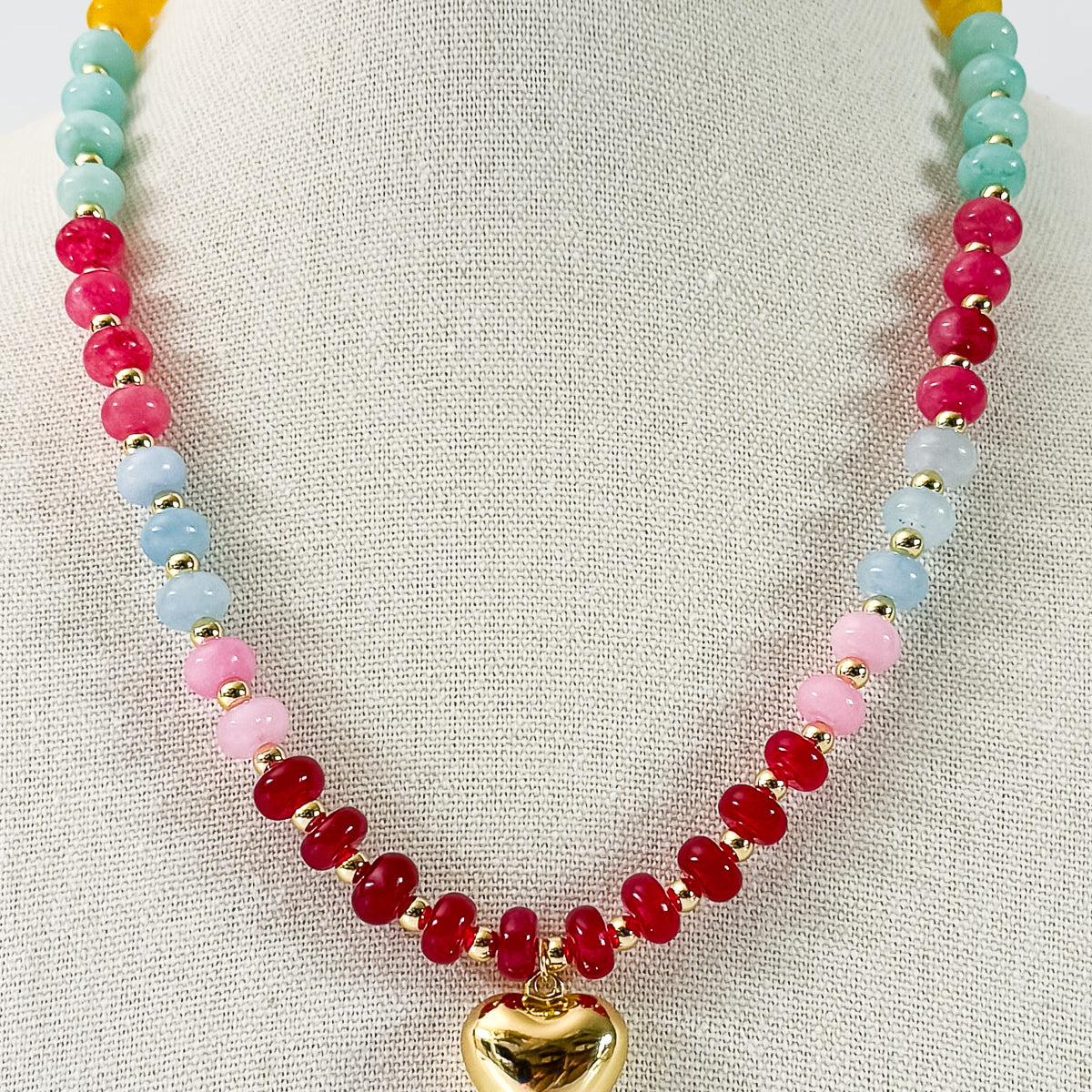 A colorful natural stone beaded necklace blending pink, blue, and yellow accents with a gold heart pendant displayed on a mannequin.