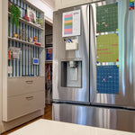 A stainless steel French-door refrigerator in a kitchen setting, with colorful monthly calendar hanging on it. To the left is a hanging shelving unit backed with striped blue-and-green wallpaper and featuring a family command and decorative items.