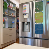 A stainless steel French-door refrigerator in a kitchen setting, with colorful monthly calendar hanging on it. To the left is a hanging shelving unit backed with striped blue-and-green wallpaper and featuring a family command and decorative items.