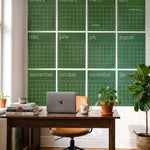 A set of twelve green monthly calendar posters arranged in a 4 x 3 grid on a white wall above a wooden desk with a laptop, plants, and office chair.