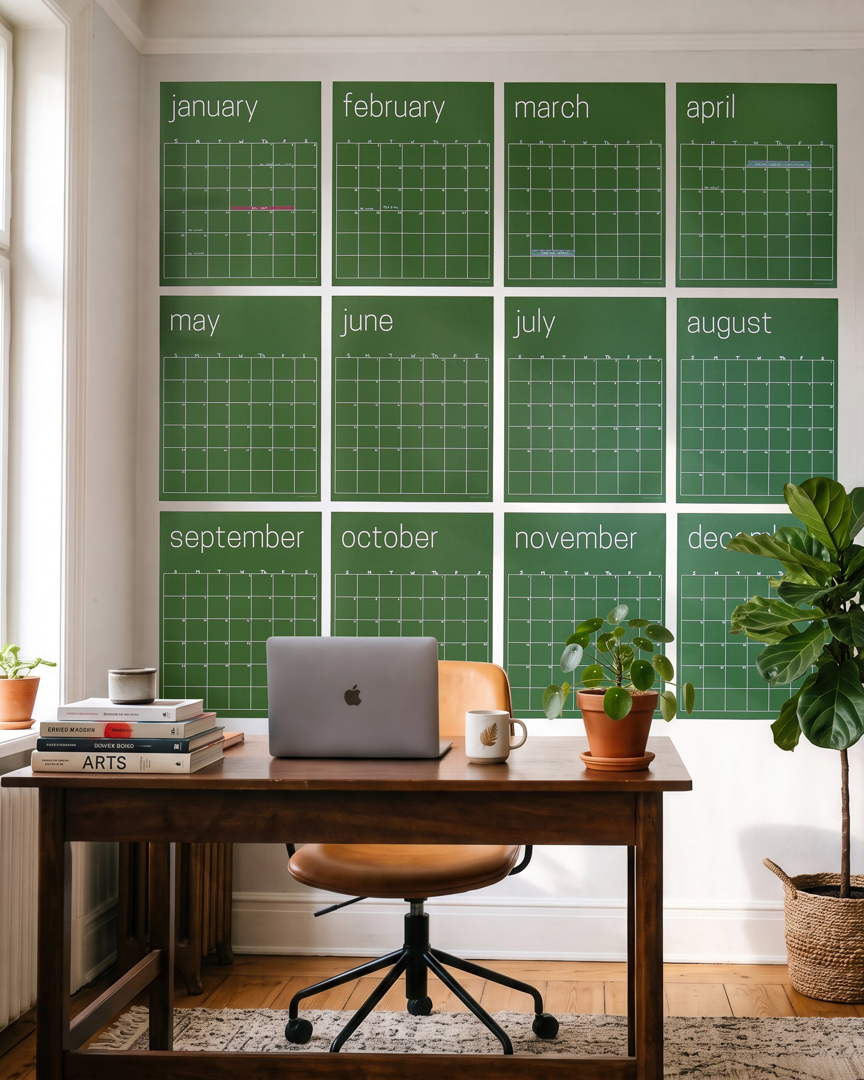 A set of twelve green monthly calendar posters arranged in a 4 x 3 grid on a white wall above a wooden desk with a laptop, plants, and office chair.