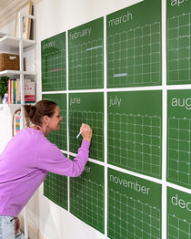 Woman writing on a large green undated, reusable wall calendar with a white chalk marker
