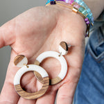 Wooden and white resin hoop earrings held in a hand with colorful beaded bracelets on her wrist