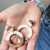 Wooden and white resin hoop earrings held in a hand with colorful beaded bracelets on her wrist