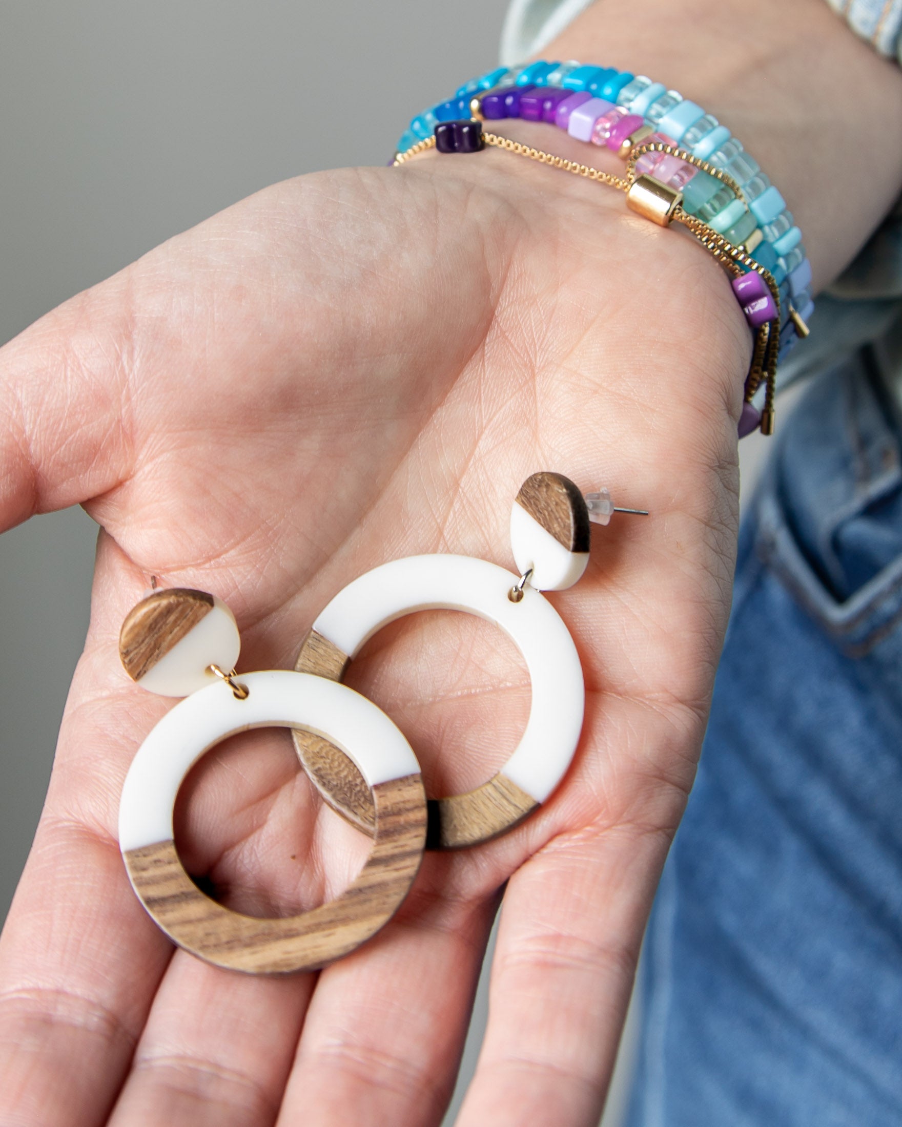 Wooden and white resin hoop earrings held in a hand with colorful beaded bracelets on her wrist