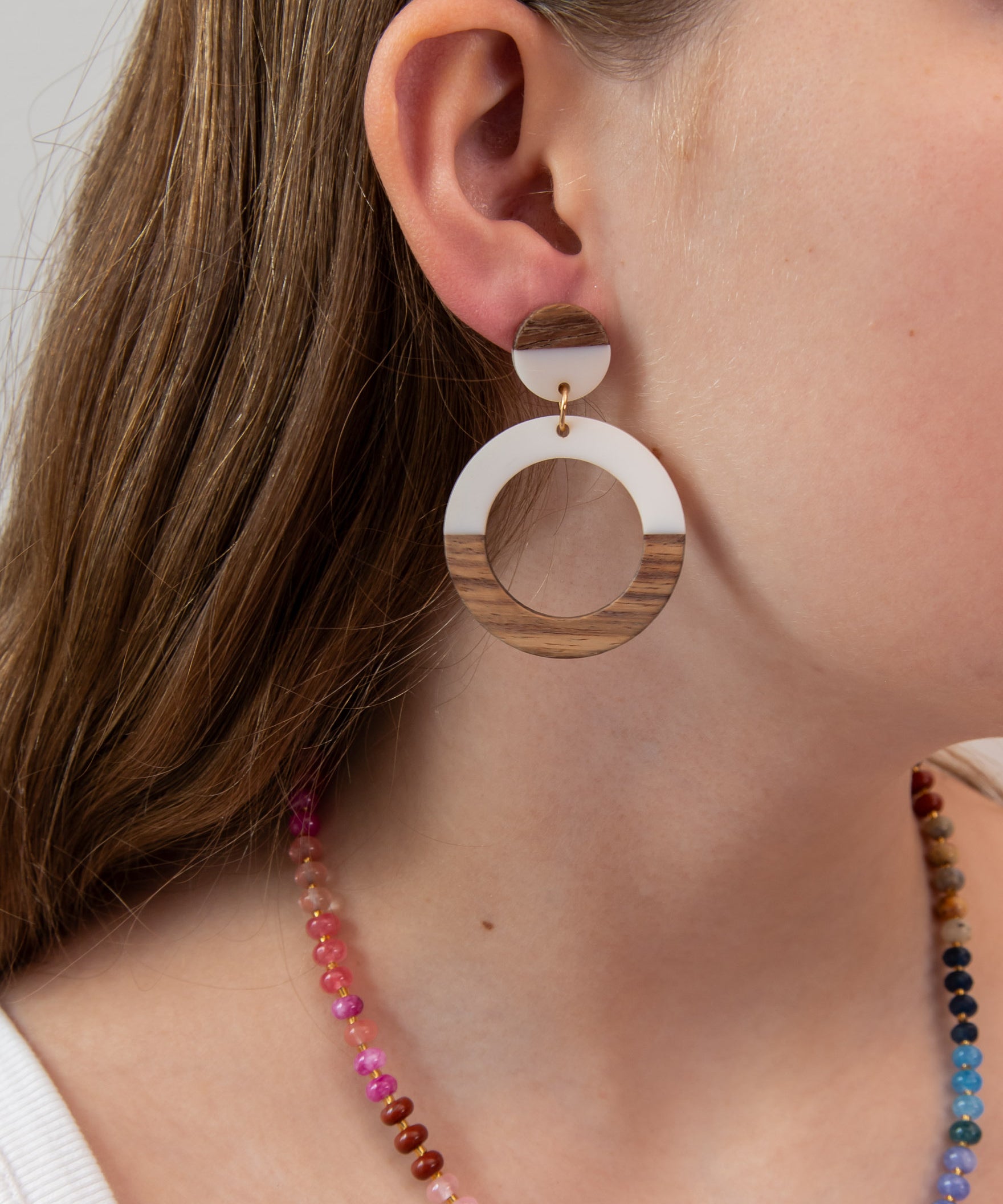 Close-up of a person wearing a white acrylic and wood hoop earring with a colorful necklace 