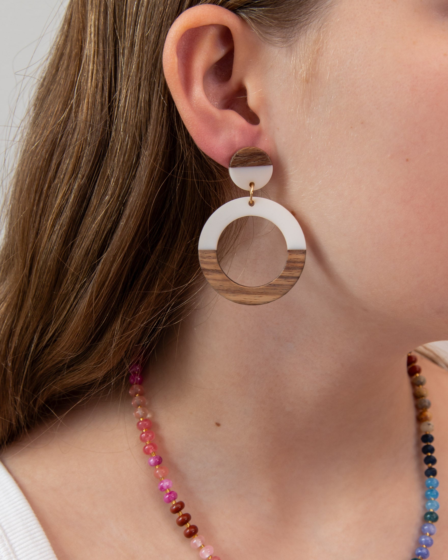 Close-up of a person wearing a white acrylic and wood hoop earring with a colorful necklace 