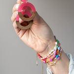 Hand holding a pair of pink and wooden hoop earrings with several colorful bracelets on her wrist 