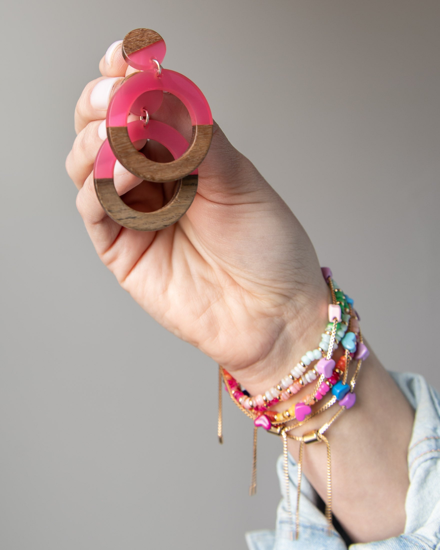 Hand holding a pair of pink and wooden hoop earrings with several colorful bracelets on her wrist 
