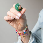 Hand holding two green acrylic hoop earrings with colorful bracelets on a neutral background