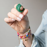 Hand holding two green acrylic hoop earrings with colorful bracelets on a neutral background