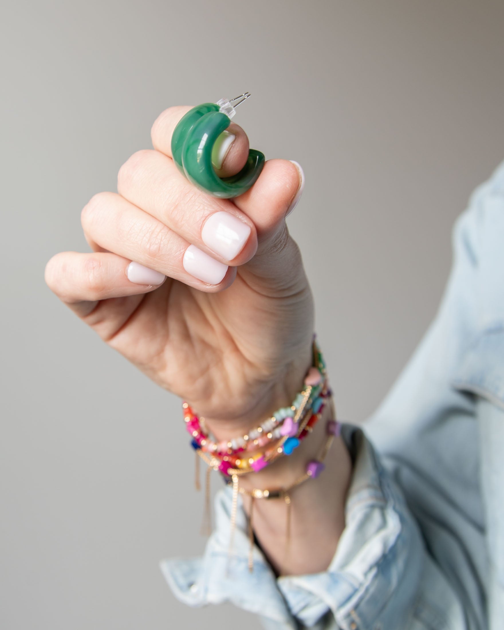 Hand holding two green acrylic hoop earrings with colorful bracelets on a neutral background