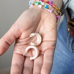 Hand holding two acrylic hoop earrings in cream with colorful bracelet on a neutral background