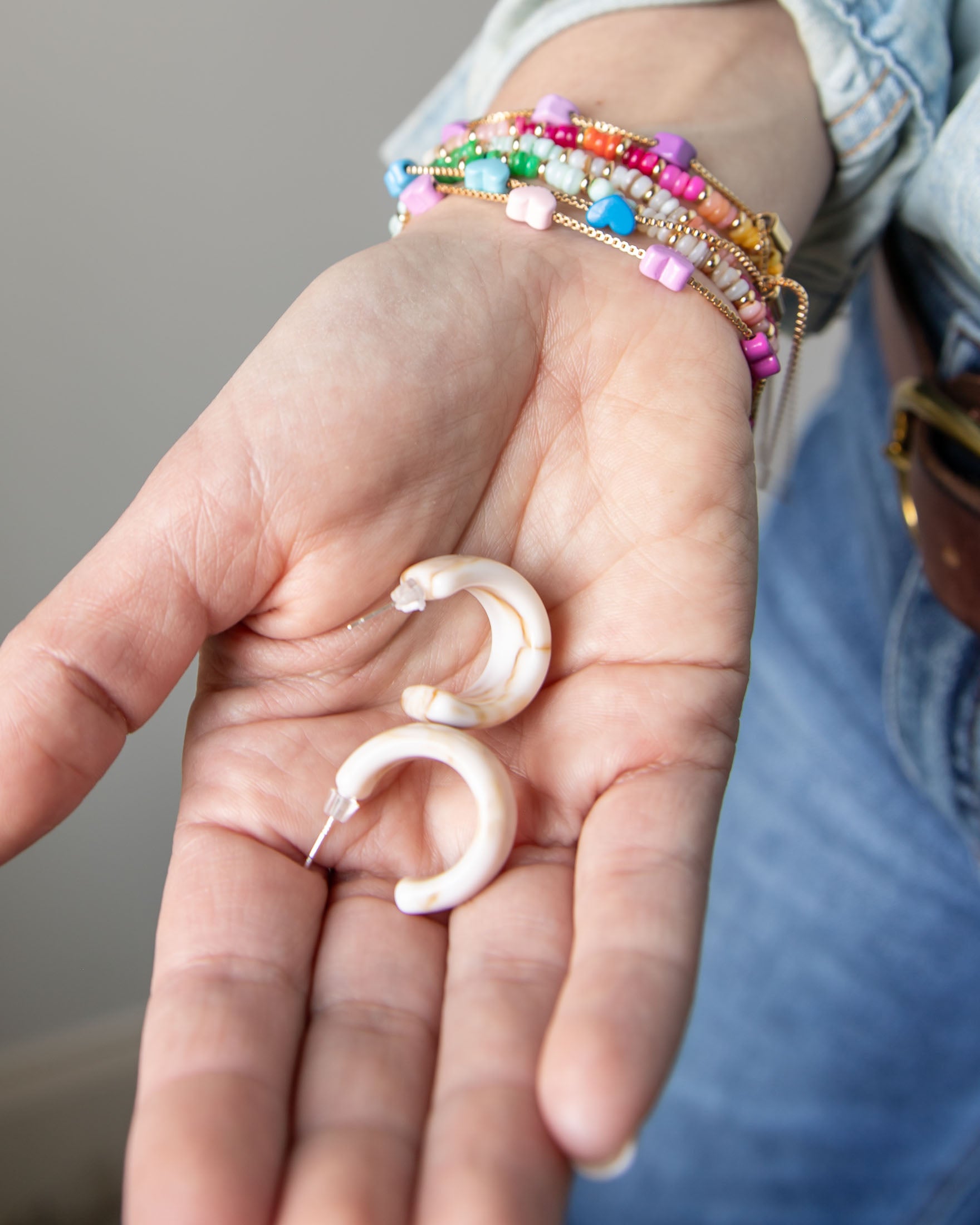 Hand holding two acrylic hoop earrings in cream with colorful bracelet on a neutral background
