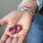 Purple acrylic, crescent-shaped earrings held in a hand with colorful bracelets on the wrist.  