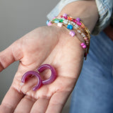 Purple acrylic, crescent-shaped earrings held in a hand with colorful bracelets on the wrist.  