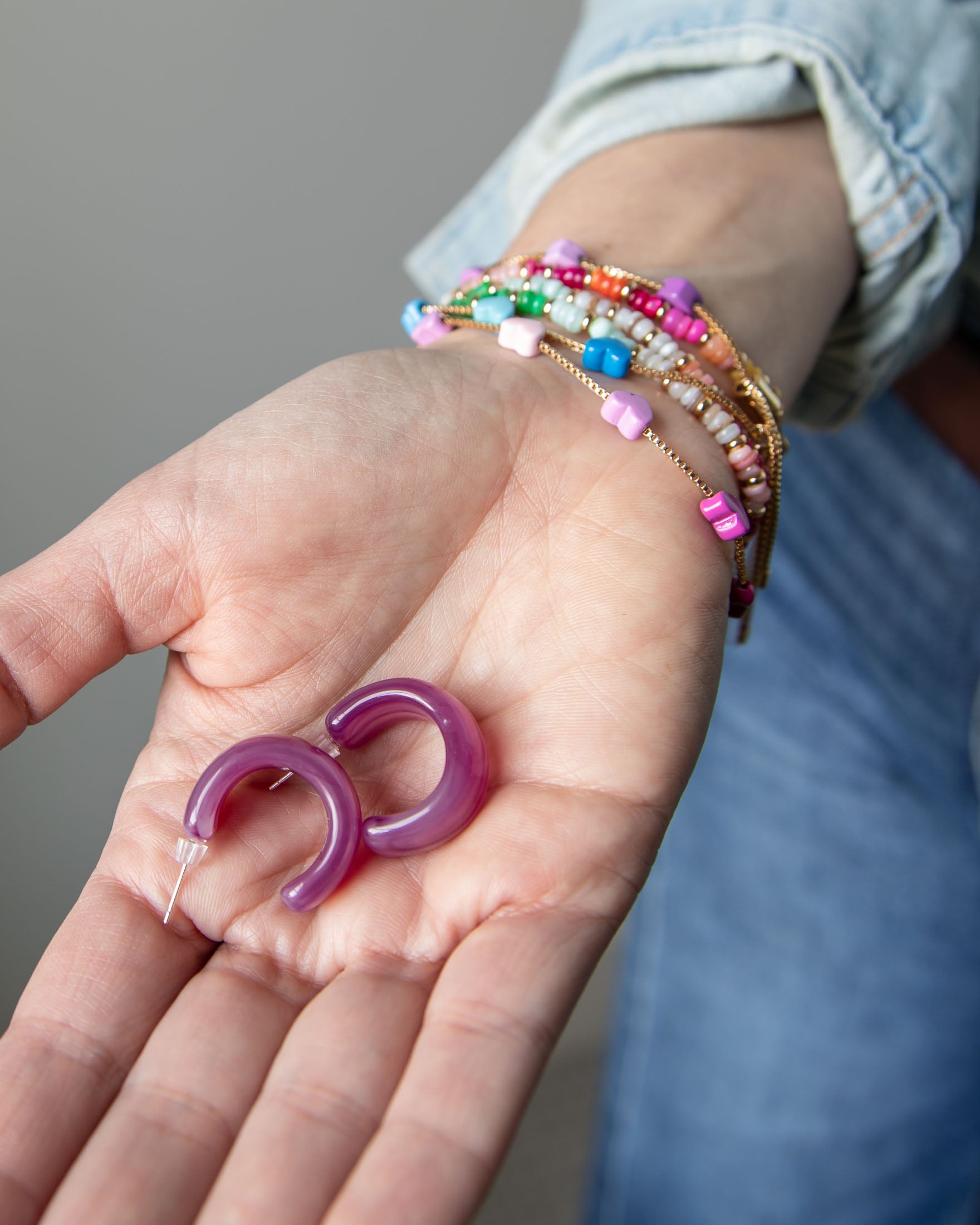 Purple acrylic, crescent-shaped earrings held in a hand with colorful bracelets on the wrist.  