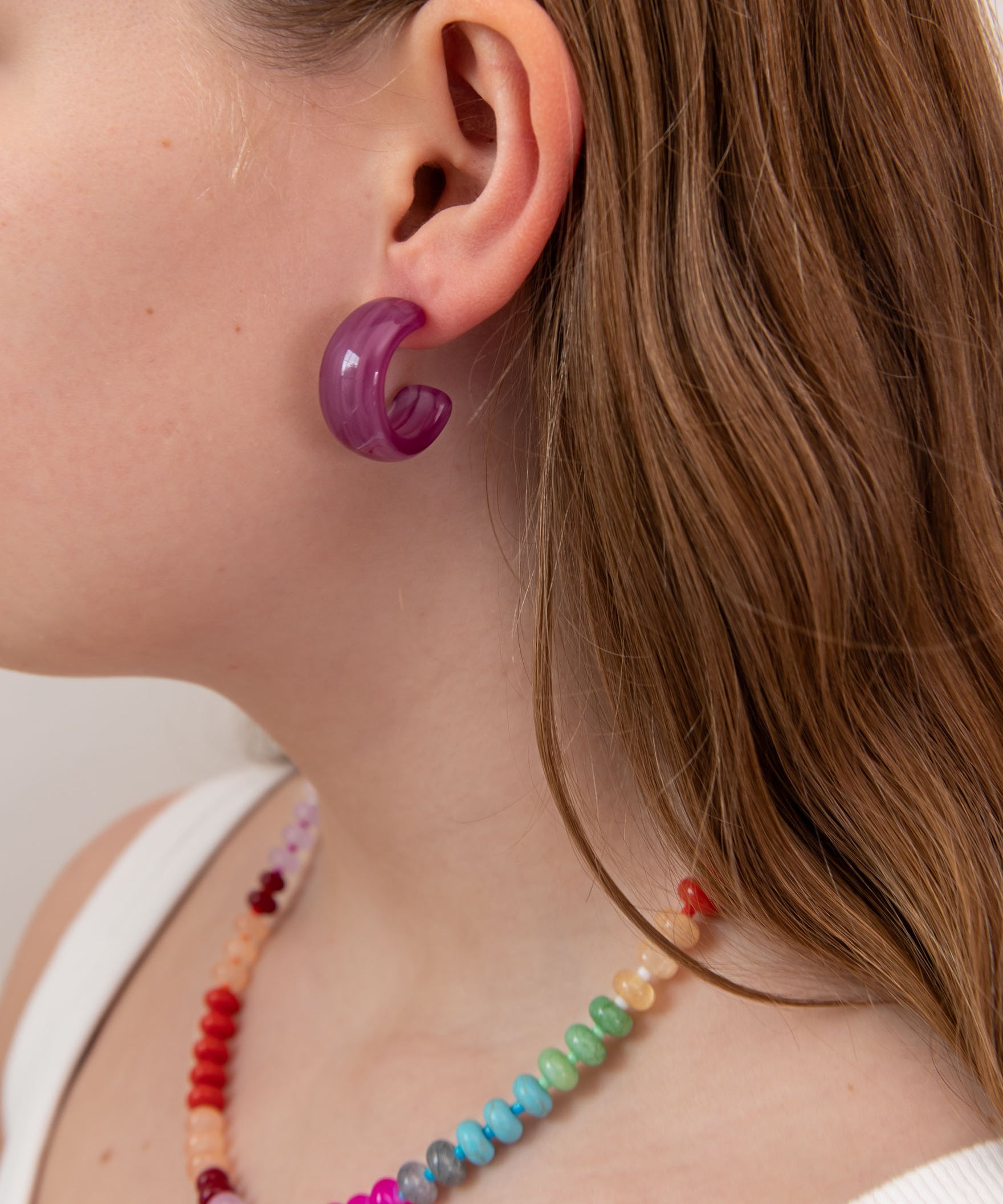 Close-up of a person wearing a purple acrylic hoop earring and colorful beaded necklace.