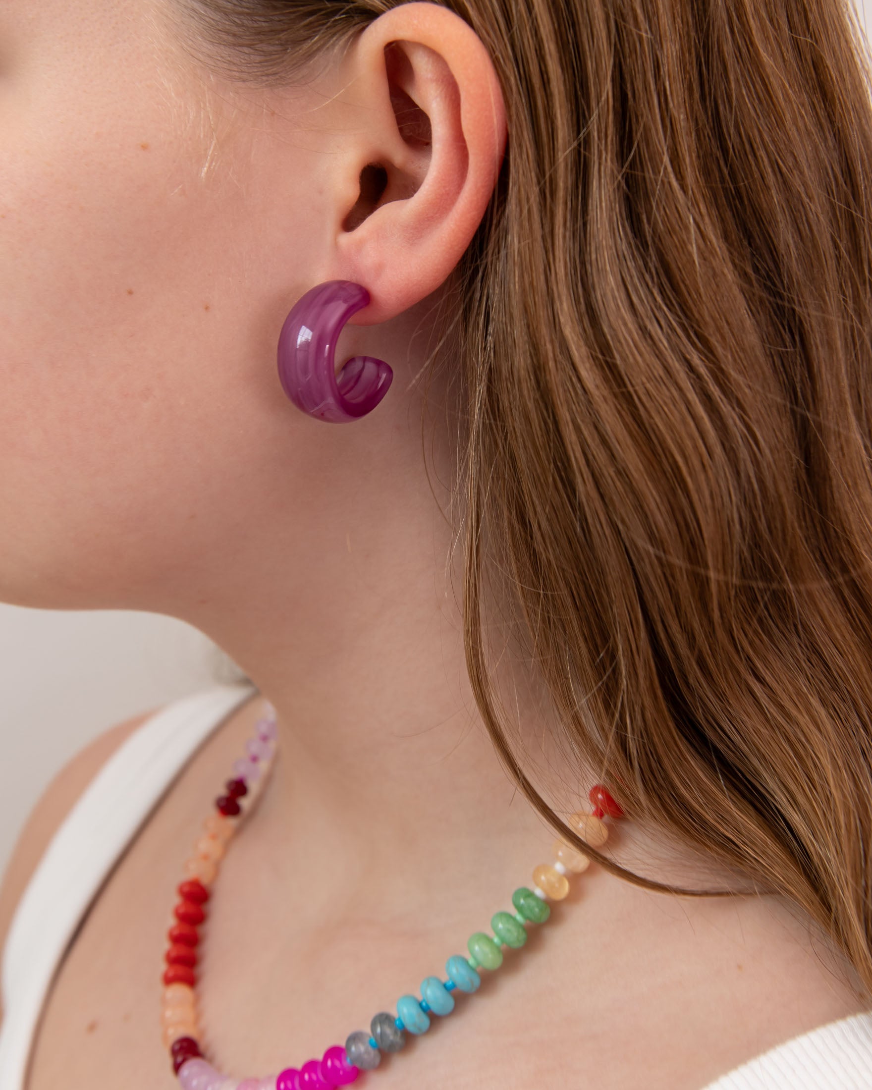 Close-up of a person wearing a purple acrylic hoop earring and colorful beaded necklace.