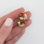 Close up of gold and colorful striped huggie earrings held between fingers by Kaleidoscope Living