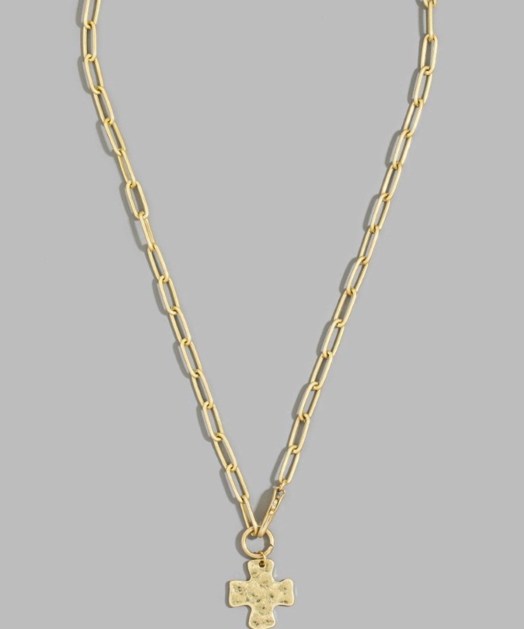 A gold paperclip chain necklace with a small gold cross pendant, displayed against a solid gray background.