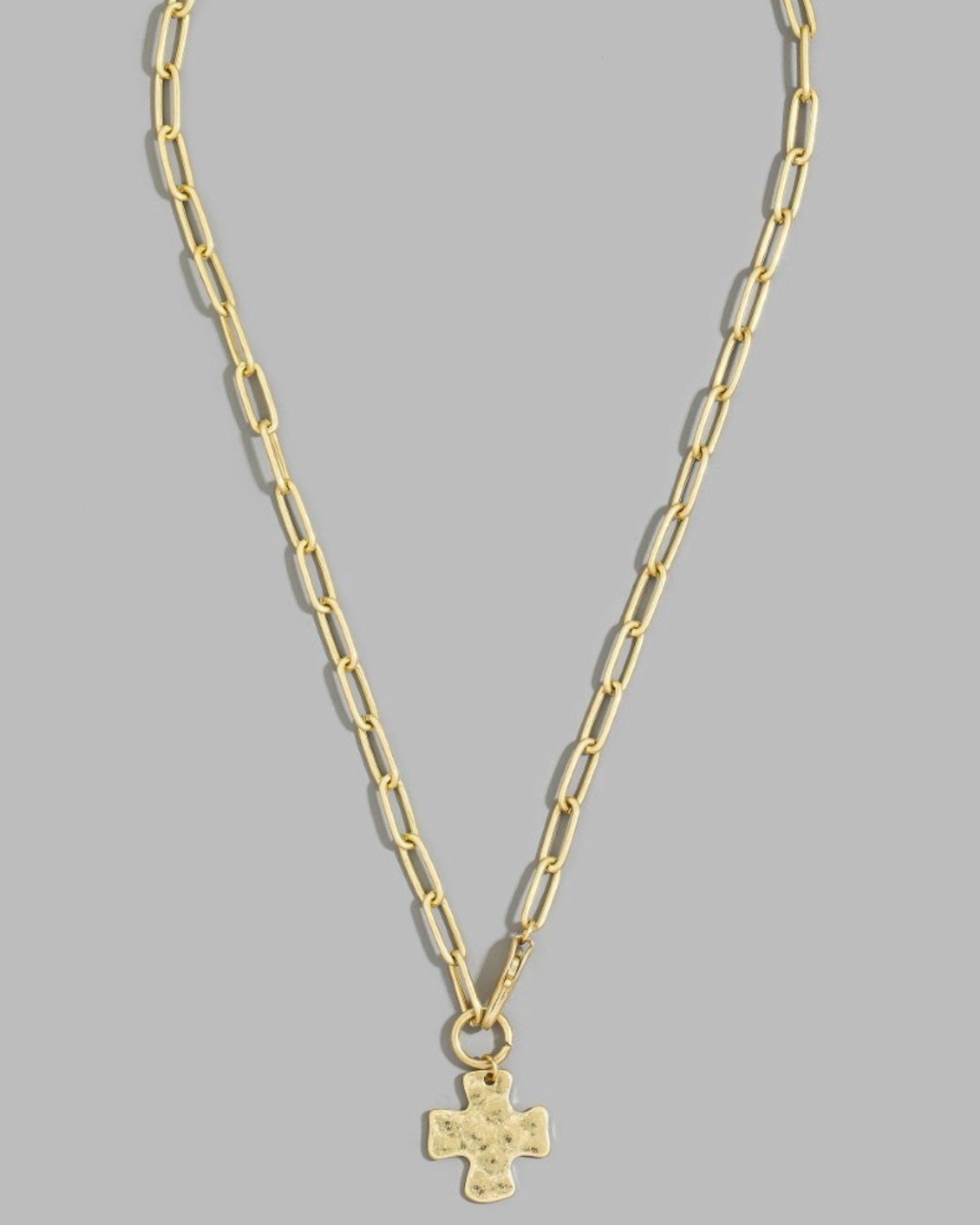A gold paperclip chain necklace with a small gold cross pendant, displayed against a solid gray background.