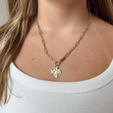 A close-up of a model wearing a gold paperclip chain necklace with a small hammered cross pendant, paired with a white ribbed tank top.