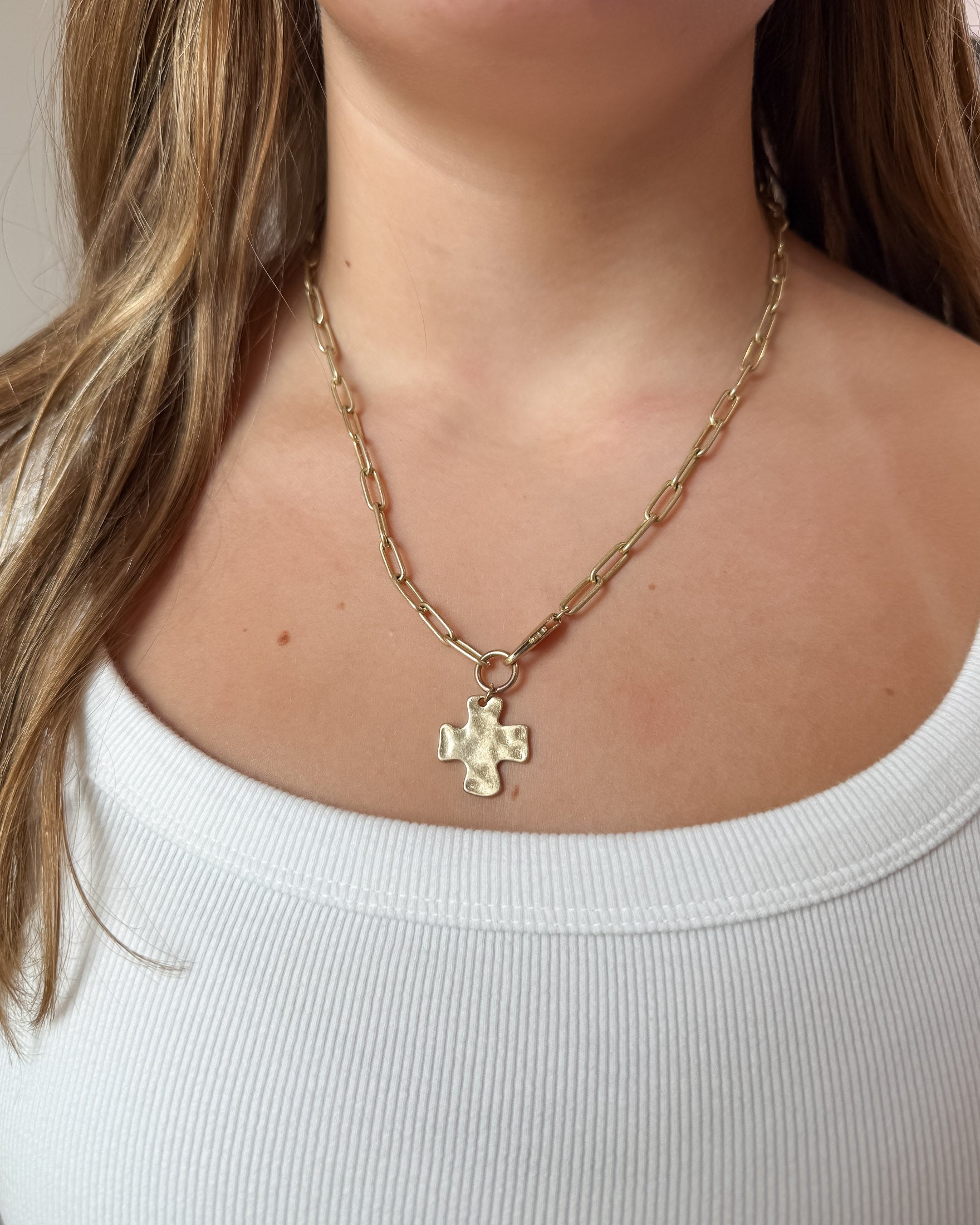 A close-up of a model wearing a gold paperclip chain necklace with a small hammered cross pendant, paired with a white ribbed tank top.