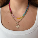 A close-up of a woman wearing a layered necklace set: one gold paperclip chain necklace with a hammered cross pendant, and colorful gemstone necklace with pink, yellow, green, blue, and purple beads.