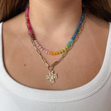 A close-up of a woman wearing a layered necklace set: one gold paperclip chain necklace with a hammered cross pendant, and colorful gemstone necklace with pink, yellow, green, blue, and purple beads.