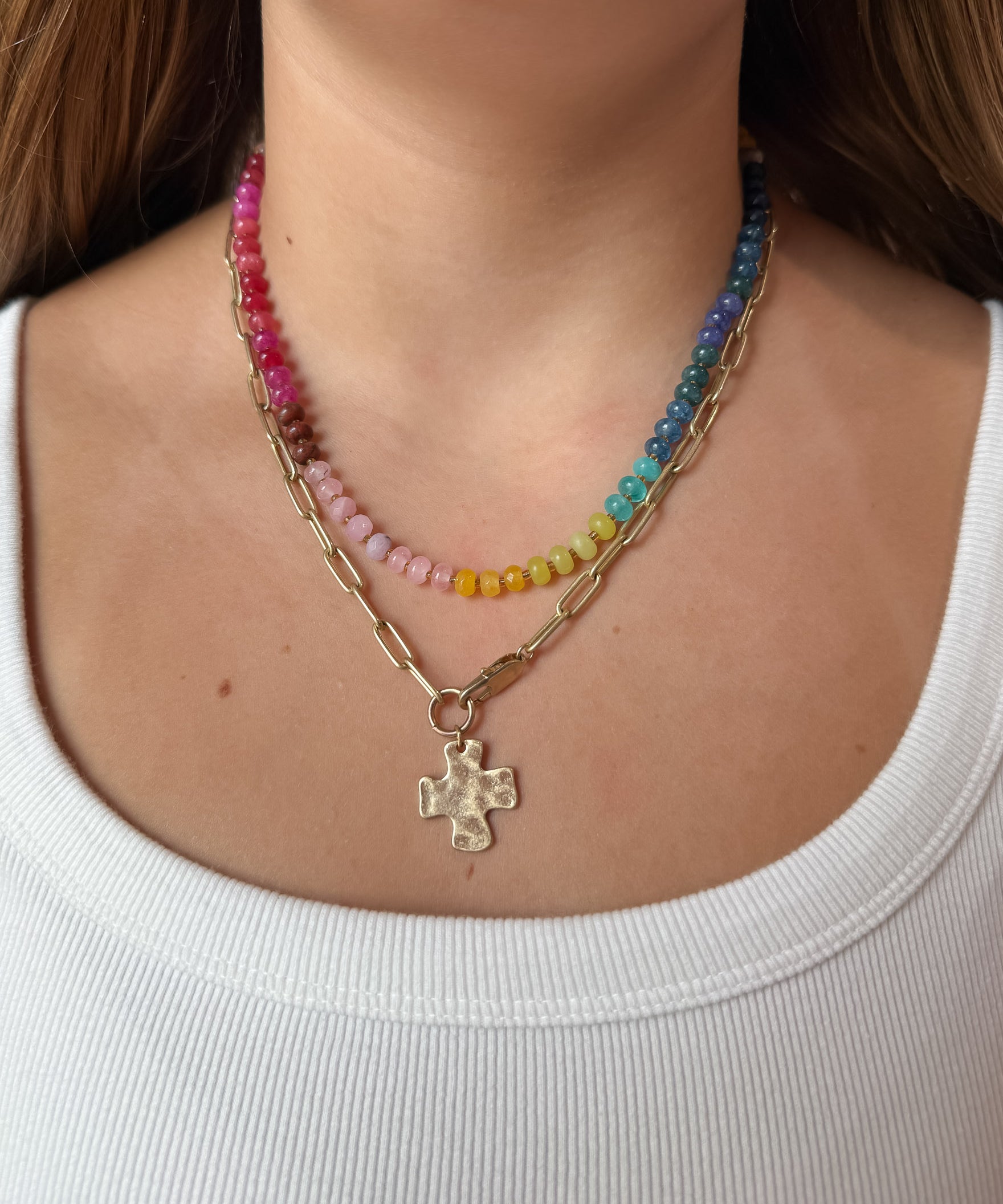 A close-up of a woman wearing a layered necklace set: one gold paperclip chain necklace with a hammered cross pendant, and colorful gemstone necklace with pink, yellow, green, blue, and purple beads.