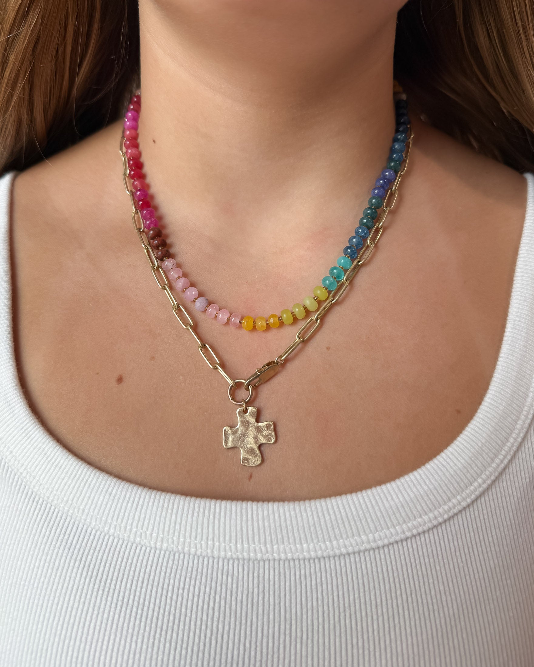 A close-up of a woman wearing a layered necklace set: one gold paperclip chain necklace with a hammered cross pendant, and colorful gemstone necklace with pink, yellow, green, blue, and purple beads.