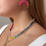 Close-up of a woman wearing layered necklaces and large pink and wood acrylic earrings. The necklaces include a gold paperclip chain with a cross pendant and a colorful beaded gemstone necklace. 