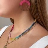 Close-up of a woman wearing layered necklaces and large pink and wood acrylic earrings. The necklaces include a gold paperclip chain with a cross pendant and a colorful beaded gemstone necklace. 