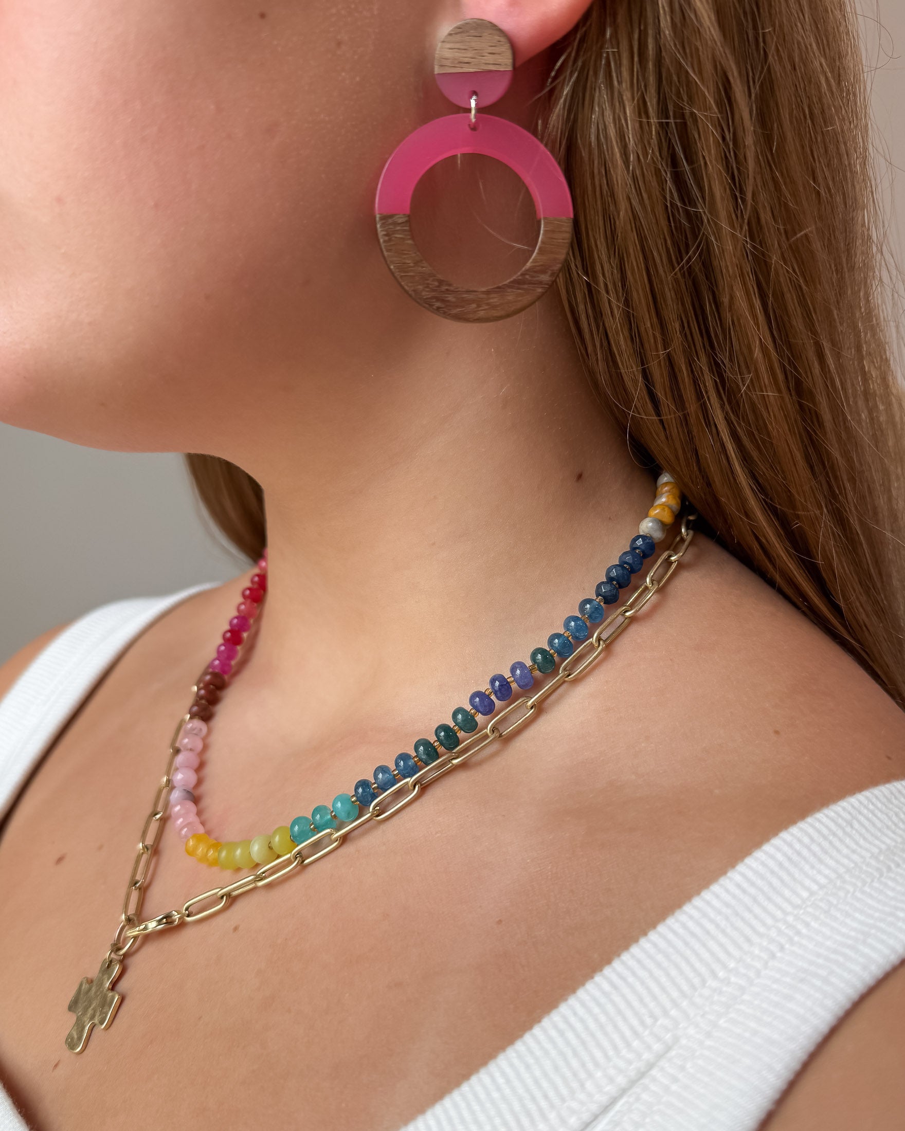Close-up of a woman wearing layered necklaces and large pink and wood acrylic earrings. The necklaces include a gold paperclip chain with a cross pendant and a colorful beaded gemstone necklace. 