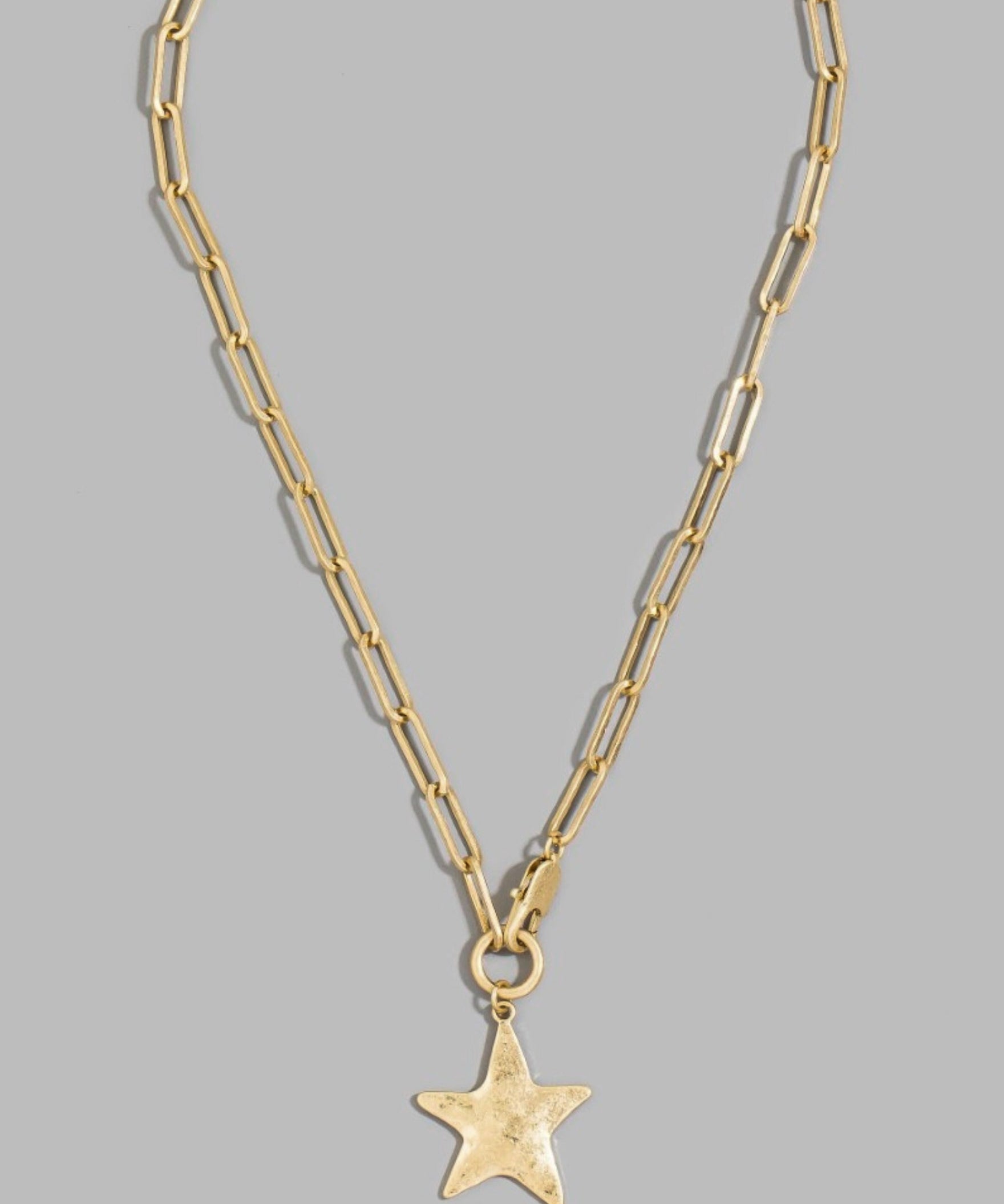 A gold paperclip chain necklace with a textured star-shaped pendant, displayed against a solid gray background.