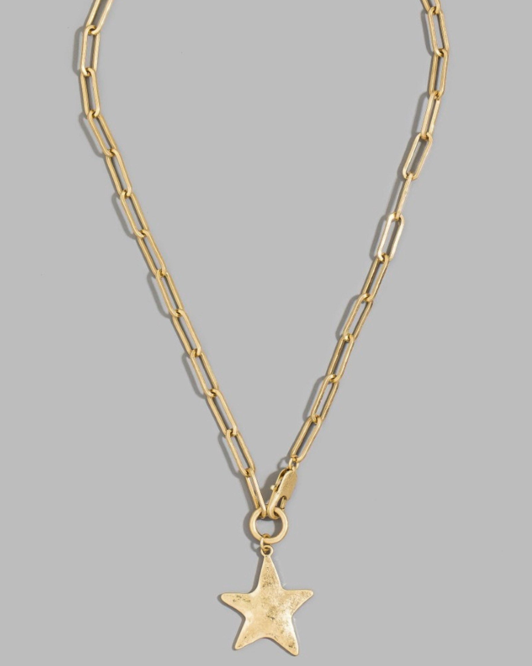 A gold paperclip chain necklace with a textured star-shaped pendant, displayed against a solid gray background.
