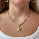 A close-up of a model wearing a gold paperclip chain necklace with a textured star pendant, paired with a white ribbed top.