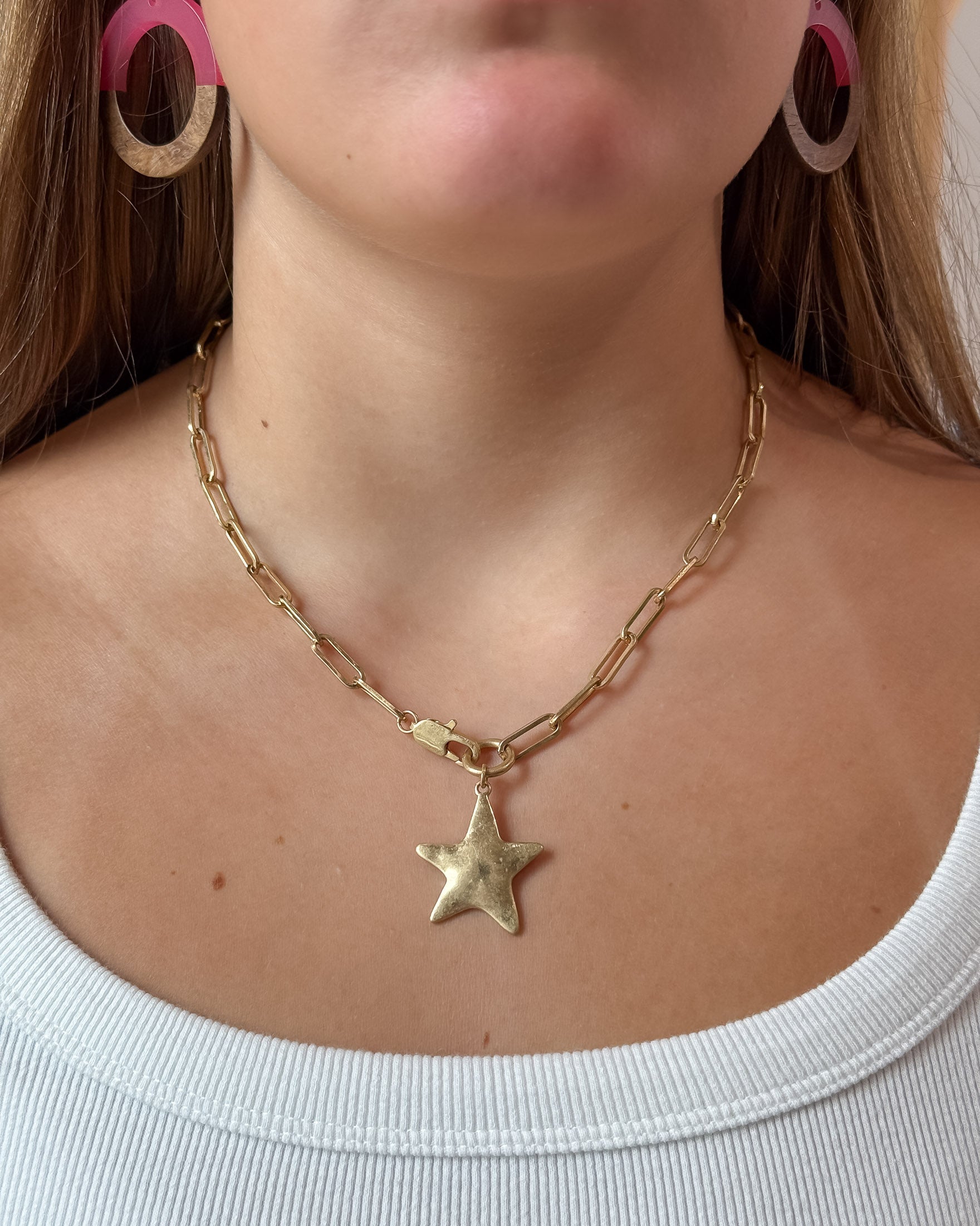 A close-up of a model wearing a gold paperclip chain necklace with a textured star pendant, paired with a white ribbed top.